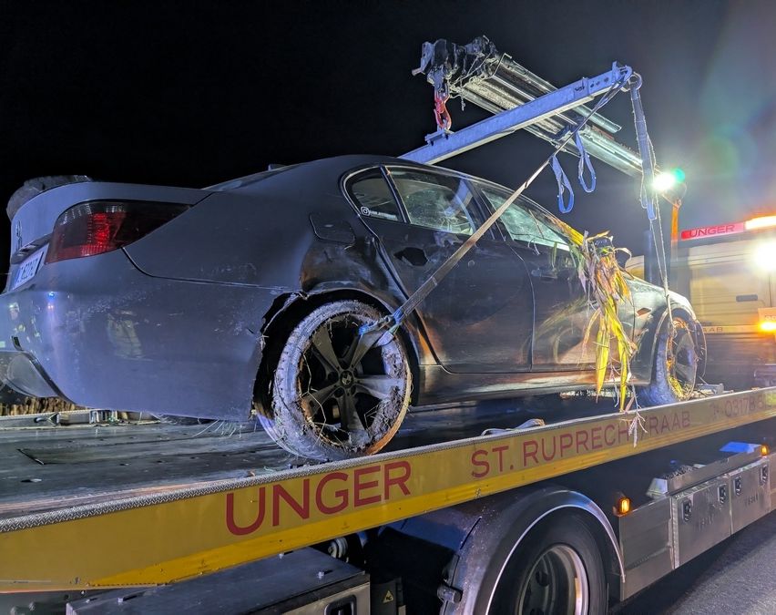 Ein beschädigtes schwarzes Auto wird von einem gelben Lastwagen mit der Aufschrift Unger abgeschleppt. Das Auto hat ein kaputtes Rad und ist mit Seilen gesichert. Die Szene spielt sich bei Nacht ab.