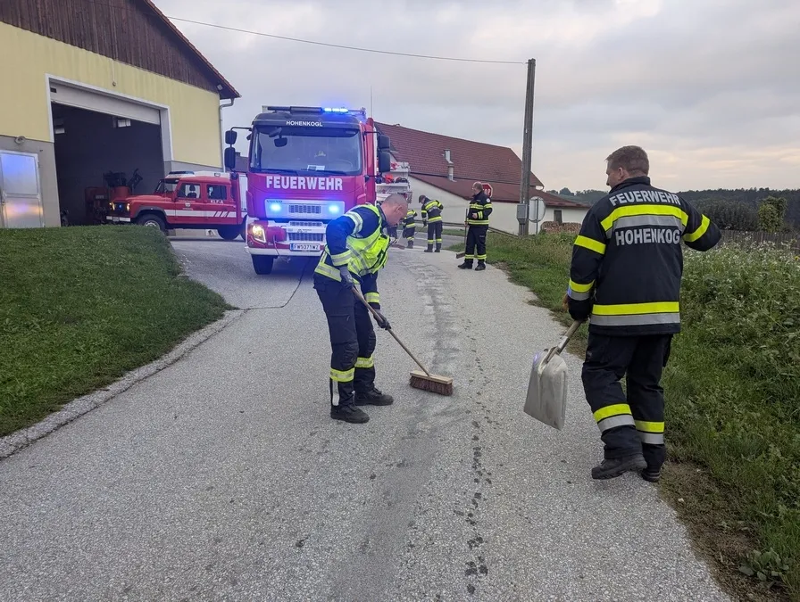 Feuerwehrleute reinigen die Straße mit einem Besen und einer Schaufel. Im Hintergrund steht ein roter Feuerwehrwagen.