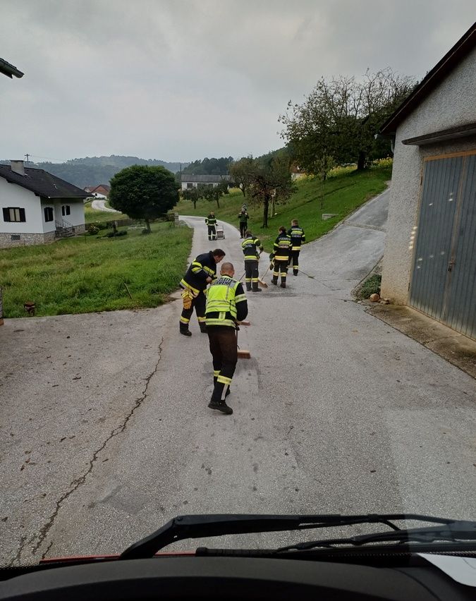 Eine Gruppe von Feuerwehrleuten in gelben und schwarzen Uniformen geht eine Straße entlang. Einige halten Besen. Im Hintergrund ein Haus und ein grünes Feld.