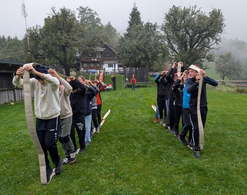 Eine Gruppe von Kindern übt das Balancieren auf Seilen in einem Grasbereich, mit einem Haus und Bäumen im Hintergrund.