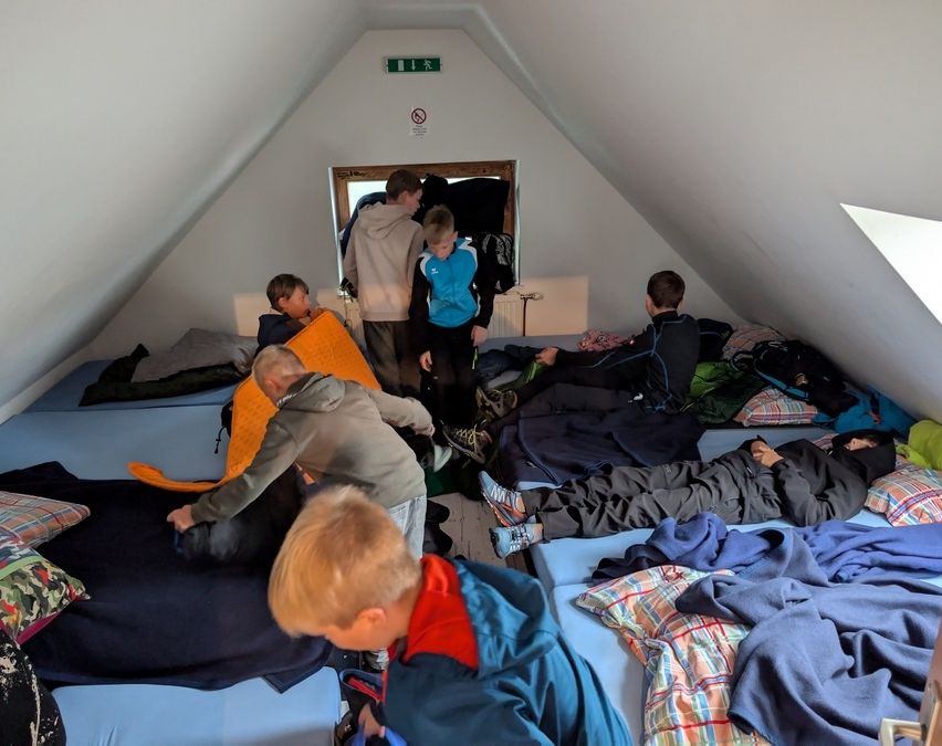 Eine Gruppe von Kindern bereitet in einem kleinen Dachzimmer ihre Betten vor. Einige ordnen Decken und Kissen, während andere bereits liegen. An der Wand befindet sich ein Schild.