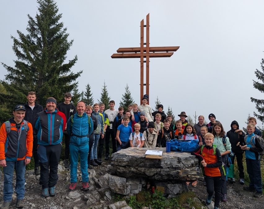 Eine Gruppe von Menschen ist an einem Berggipfel versammelt und posiert für ein Foto vor einem Holzkreuz und einem großen Stein. Sie tragen verschiedene Jacken und Schuhe.