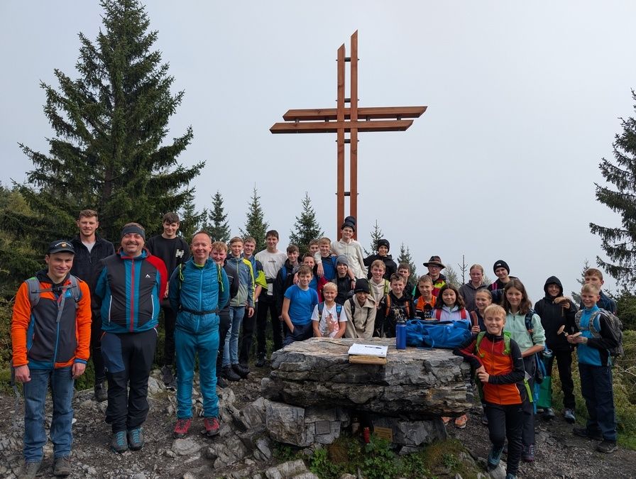 Eine Gruppe von Menschen ist an einem Berggipfel versammelt und posiert für ein Foto vor einem Holzkreuz und einem großen Stein. Sie tragen verschiedene Jacken und Schuhe.