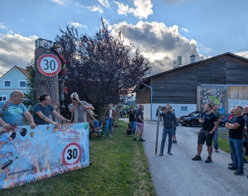 Menschen versammeln sich vor einem Gebäude mit einem Schild, das auf eine 30 km/h Geschwindigkeitsbegrenzung hinweist. Ein Banner zeigt '30', und ein Mann klettert mit einem Schild auf einen Baum. Andere stehen herum, einige halten Luftballons.