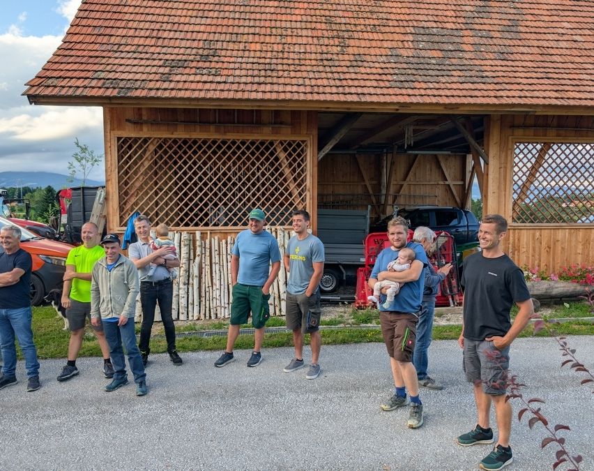 Eine Gruppe von Männern, einige mit Babys, steht vor einem roten Schuppen. Sie tragen Freizeitkleidung und Sneakers. Hinter ihnen steht ein Pickup-Truck.
