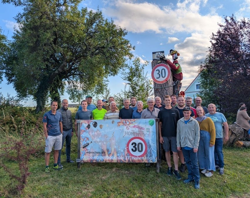 Eine Gruppe von Menschen steht vor einem Schild mit einem gemalten Tempolimit von 30 und einem Baum mit einer Maskottchen darauf. Sie sind alle lässig gekleidet, stehen auf Gras mit einem Gebäude im Hintergrund.