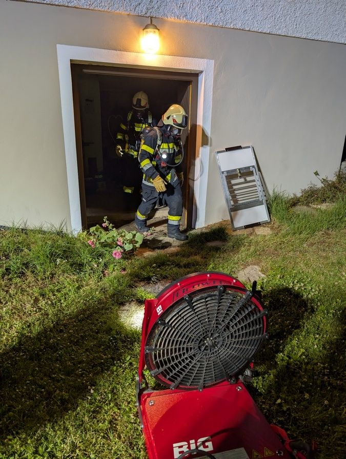 Zwei Feuerwehrleute in Schutzkleidung betreten ein Gebäude, einer von ihnen benutzt eine Taschenlampe. Ein roter Ventilator steht auf dem Rasen.