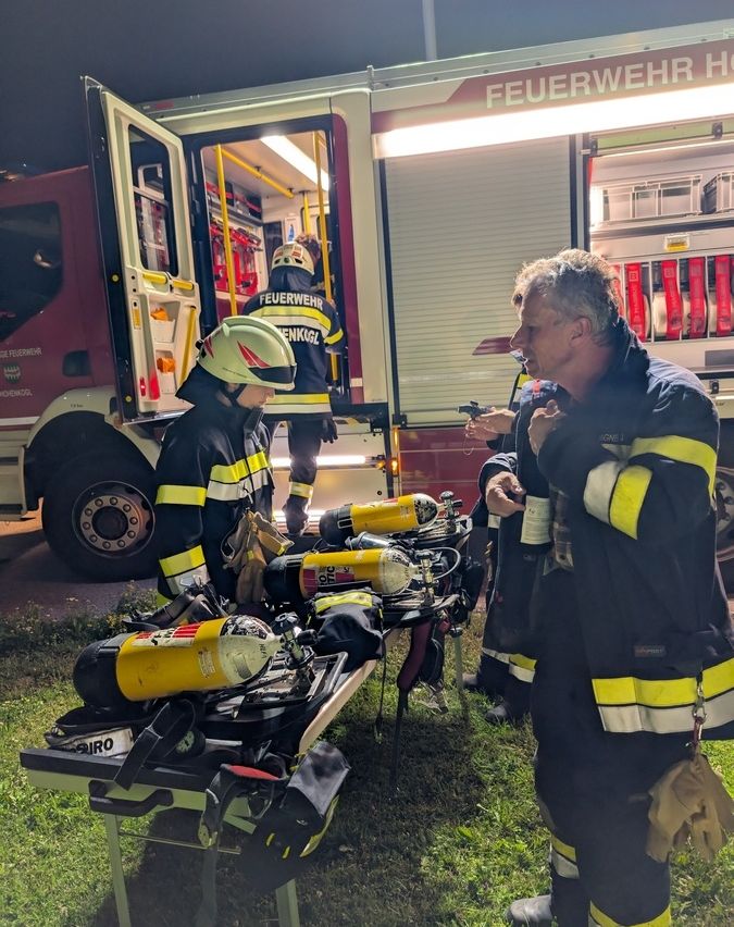 Ein Feuerwehrteam in Schutzkleidung steht neben einem Feuerwehrwagen. Ein Feuerwehrmann spricht mit einem anderen. Ausrüstung und Sauerstoffflaschen sind auf einem Tisch vor ihnen platziert.