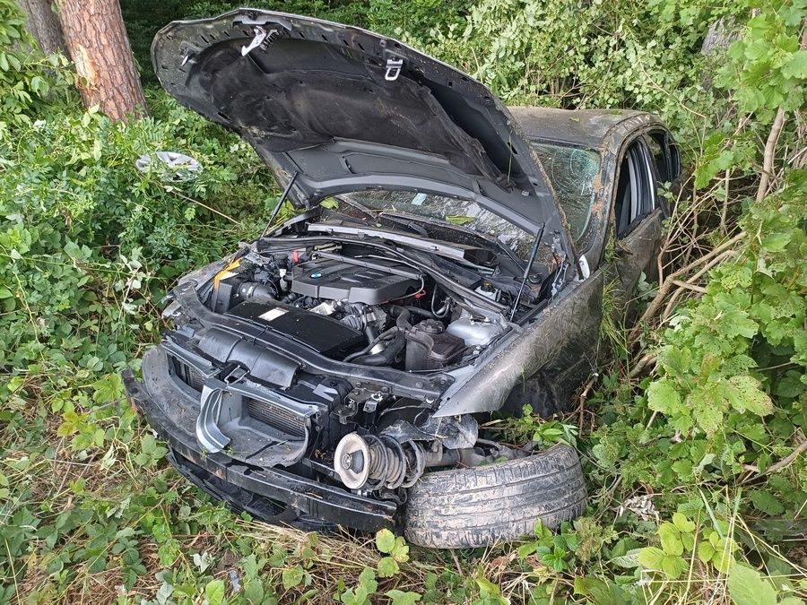 Ein beschädigtes Auto steckt in den Büschen fest, mit offenem Motorhaube, der Motor und der vordere Stoßfänger sind sichtbar.