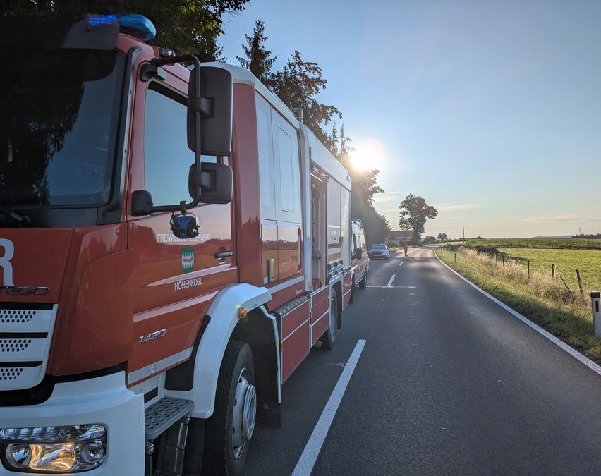 Ein roter Feuerwehrwagen ist am Straßenrand geparkt, mit seiner offenen Tür, die den Innenraum zeigt. Die Sonne geht im Hintergrund unter, und ein Auto fährt weg.