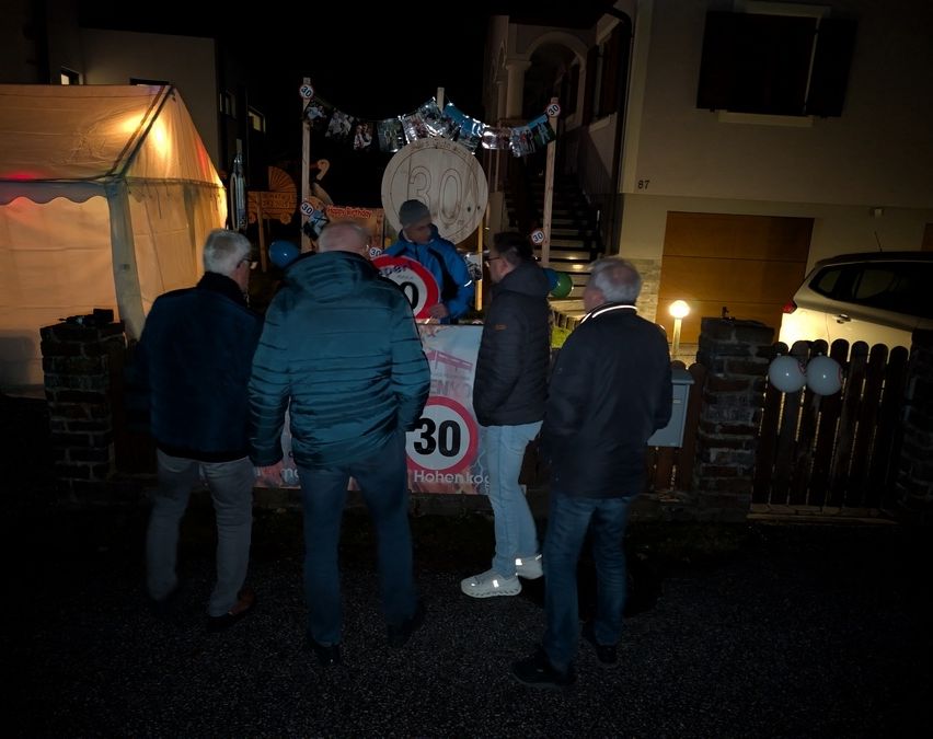 Fünf Männer stehen nachts vor einem Haus. Ein Geschwindigkeitsbegrenzungsschild mit 30 ist sichtbar. Es gibt Dekorationen und Ballons.