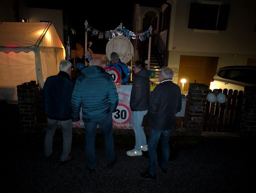 Fünf Männer stehen nachts vor einem Haus. Ein Geschwindigkeitsbegrenzungsschild mit 30 ist sichtbar. Es gibt Dekorationen und Ballons.