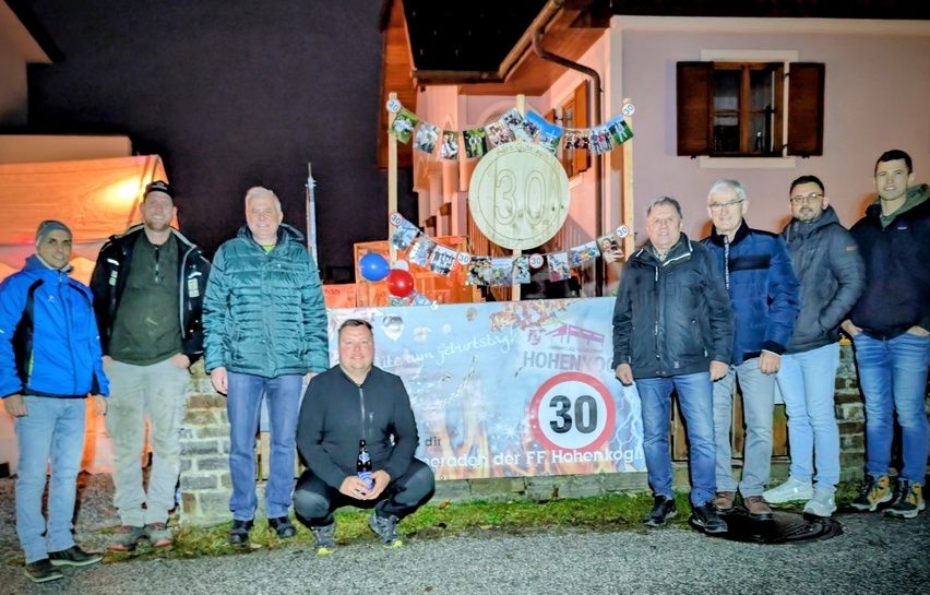 Eine Gruppe von Männern posiert für ein Foto vor einem Banner mit Bildern und einer 30er-Tafel. Sie tragen Jacken und einige halten Flaschen. Die Szene spielt sich nachts ab.