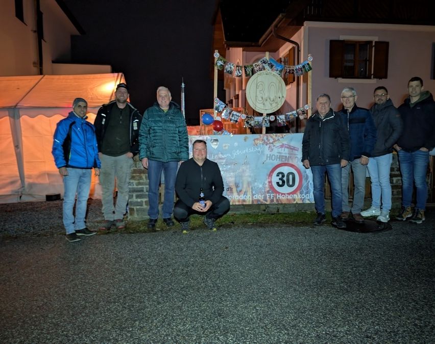 Eine Gruppe von Männern in Winterjacken steht vor einem Banner und einem Gebäude mit einem Schild, auf dem '30' steht. Dahinter befindet sich ein weißes Zelt und ein Gebäude mit einem Fenster.