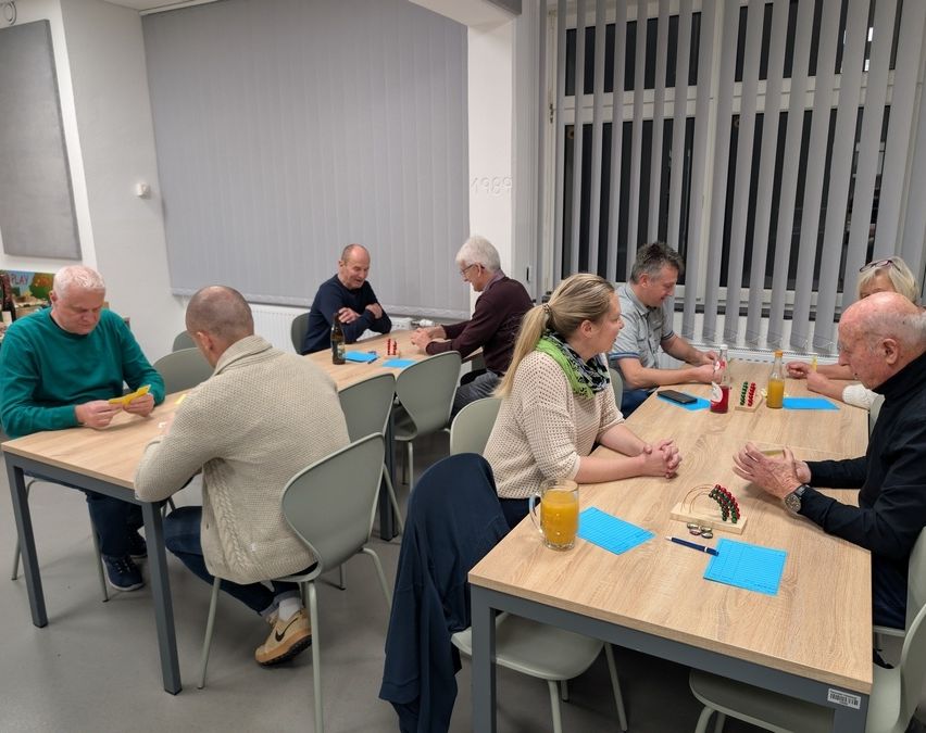 Mehrere Personen sitzen in einem Raum um einen Tisch herum und scheinen ein Spiel zu spielen. Jede Person hat ein Glas Orangensaft. Eine Person trägt eine Schürze.