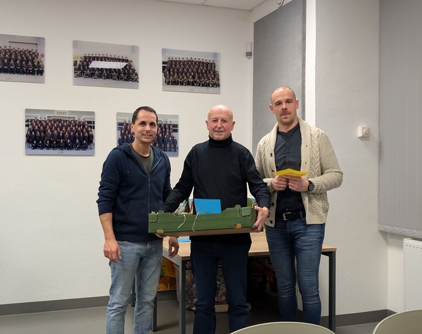 Drei Männer stehen in einem Raum, einer hält eine grüne Box. Der Mann in der Mitte lächelt und schaut auf die Box. Dahinter steht ein Tisch mit einer Tafel und Bilderrahmen an der Wand.