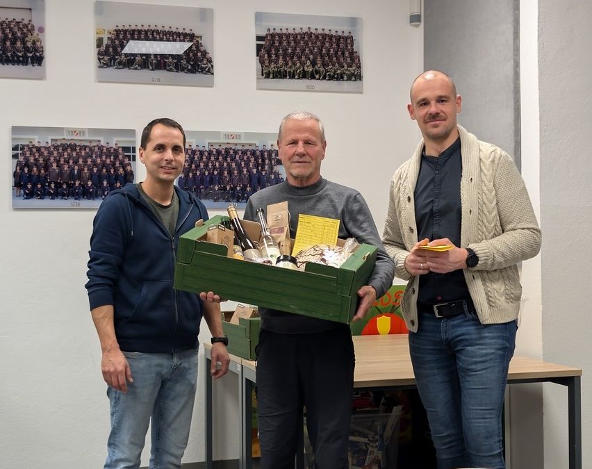 Drei Männer stehen hinter einem Tisch, zwei lächeln und einer hält eine Box mit Weinflaschen. Sie sind in einem Raum mit gerahmten Fotos an der Wand.
