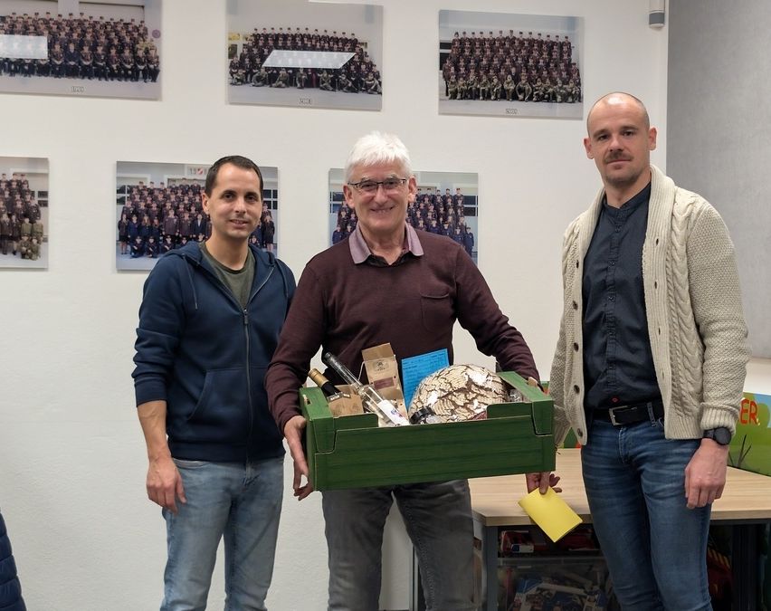 Drei Männer stehen in einem Raum mit einem Tisch dahinter, einer hält eine Box mit Gegenständen, ein anderer lächelt und der dritte hat einen ernsten Ausdruck. Sie stehen vor einer Wand mit Fotos von Gruppen von Menschen.