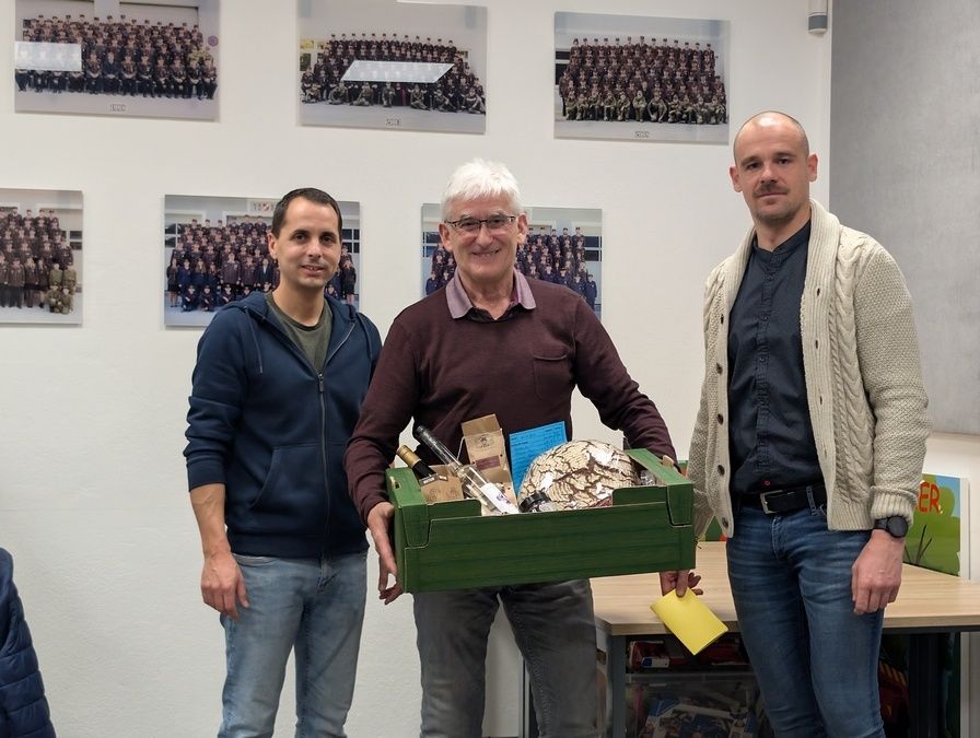 Drei Männer stehen in einem Raum mit einem Tisch dahinter, einer hält eine Box mit Gegenständen, ein anderer lächelt und der dritte hat einen ernsten Ausdruck. Sie stehen vor einer Wand mit Fotos von Gruppen von Menschen.