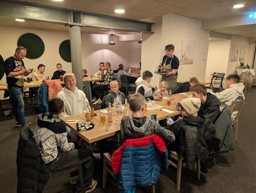 Eine Gruppe von Personen sitzt in einem Restaurant um einen langen Tisch herum, einige in Winterjacken, unterhält sich beim Trinken. Ein Mann in einem Schürzen serviert Getränke auf einem Tablett.