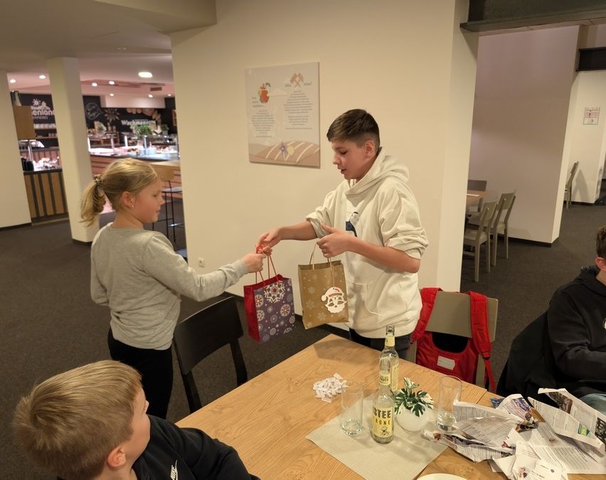 Zwei Kinder tauschen Geschenke in einem Raum mit einem Holztisch, Stühlen und einer an der Wand montierten Speisekarte aus. Ein Kind trägt eine Kapuzenjacke und hält eine Geschenktüte.