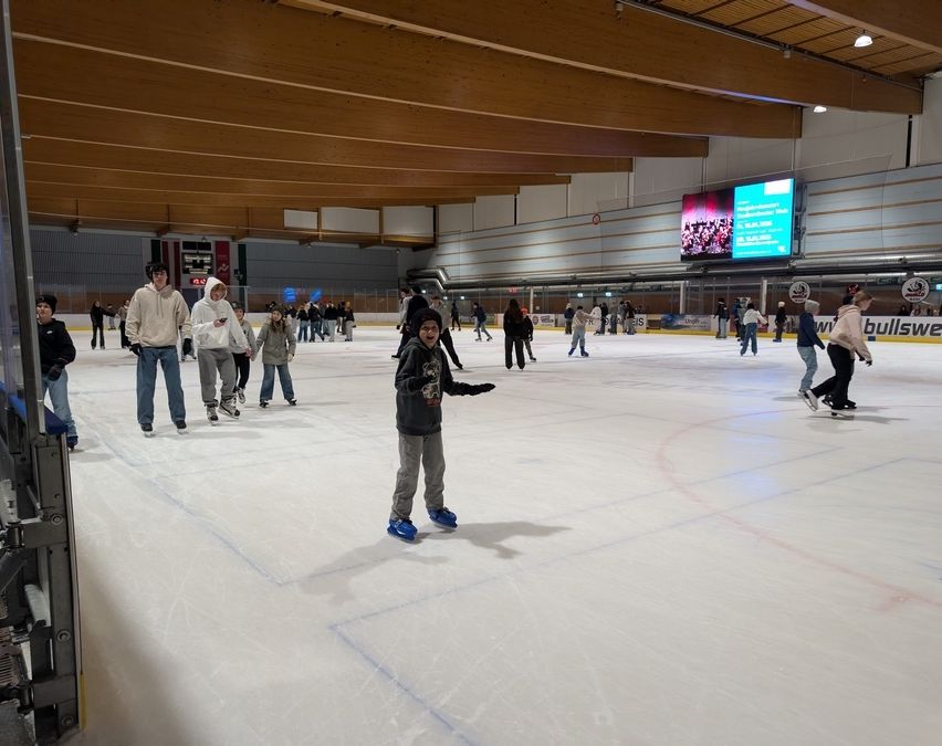 Eine überdachte Eisbahn mit mehreren Schlittschuhläufern, darunter ein Kind mit Hut und Handschuhen. An der Wand ist ein großer Monitor montiert, den einige Personen beobachten.