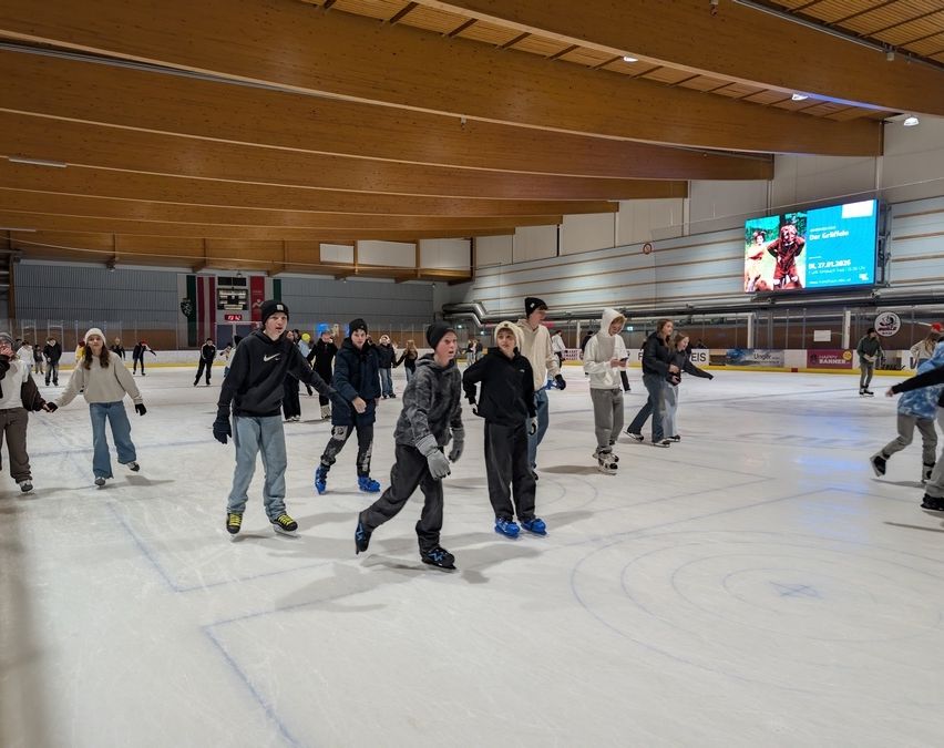 Eine Gruppe von Menschen übt das Eislaufen in einer überdachten Eisbahn. Sie tragen Winterkleidung und Schlittschuhe. Ein Monitor ist an der Wand montiert.