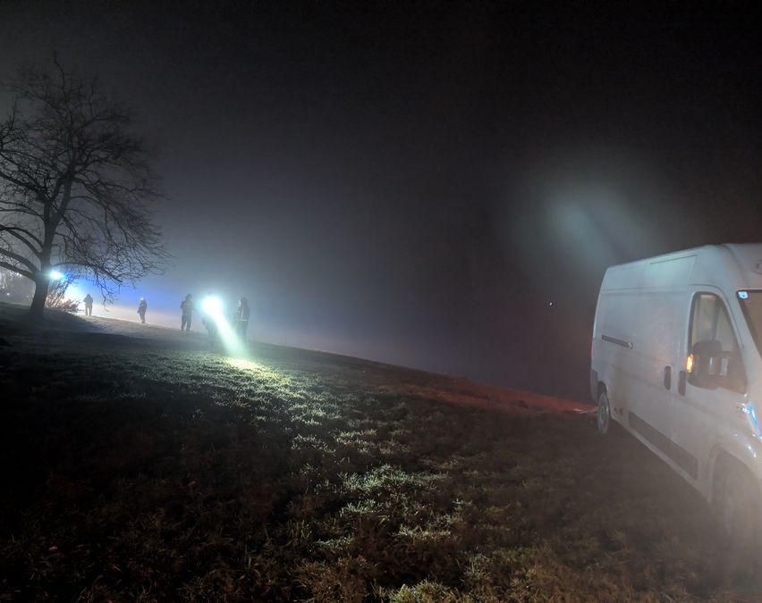 Ein weißer Lieferwagen steht mit eingeschalteten Scheinwerfern auf einem grasigen Hügel und beleuchtet eine Gruppe von Personen mit Taschenlampen.