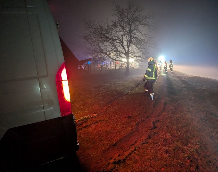 Feuerwehrleute in Schutzkleidung navigieren einen schlammigen Pfad bei Nacht in nebligen Bedingungen. Links ist das Rücklicht eines Fahrzeugs zu sehen.