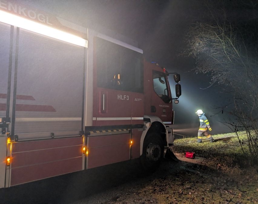 Ein Feuerwehrmann in Uniform steht neben einem roten Feuerwehrwagen mit der Aufschrift HLF3 auf der Seite, unter einem hellen Licht.