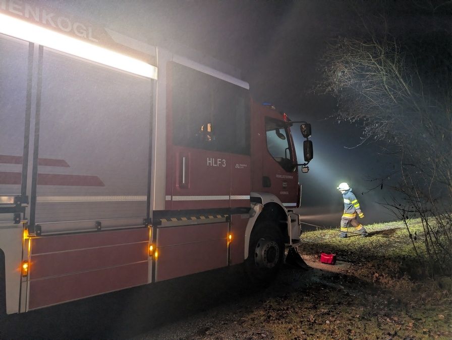 Ein Feuerwehrmann in Uniform steht neben einem roten Feuerwehrwagen mit der Aufschrift HLF3 auf der Seite, unter einem hellen Licht.