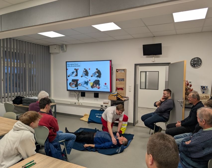 Eine Gruppe von Menschen sitzt in einem Klassenzimmer und beobachtet eine CPR-Demonstration. Ein Mann auf einer blauen Matte wird von einem anderen Mann in einer reflektierenden Weste unterstützt.