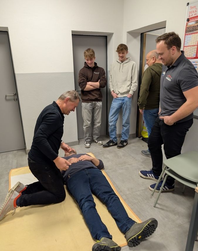 Ein Mann demonstriert CPR an einem Manikins in einem Raum, während mehrere Leute zusehen. Ein Mann kniet, ein anderer steht und zwei Männer stehen im Hintergrund und beobachten.