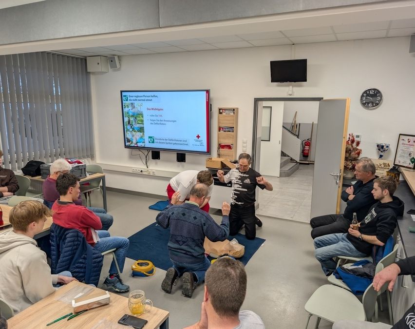 Eine Gruppe von Menschen ist in einem Raum, wahrscheinlich einem Klassenzimmer, und lernt Erste Hilfe. Ein Mann kniet auf einem blauen Teppich und demonstriert CPR an einer Puppe. Ein Monitor ist an der Wand angebracht.