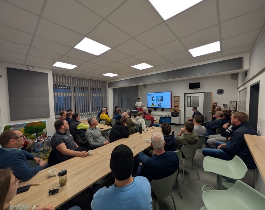 Eine Gruppe von Menschen ist in einem Raum für eine Präsentation versammelt. Ein Mann steht vorne und präsentiert an einer Projektionsleinwand. Das Publikum sitzt in Stühlen um Tische herum, und einige haben Getränke auf den Tischen. Der Raum hat ein modernes Design mit großen Fenstern.