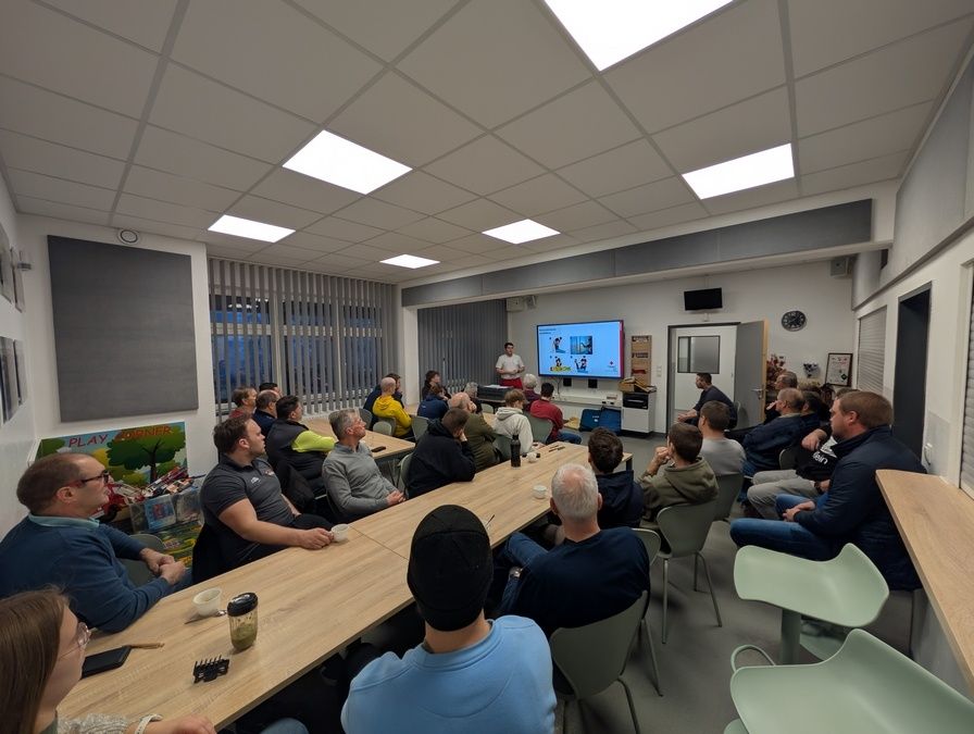 Eine Gruppe von Menschen ist in einem Raum für eine Präsentation versammelt. Ein Mann steht vorne und präsentiert an einer Projektionsleinwand. Das Publikum sitzt in Stühlen um Tische herum, und einige haben Getränke auf den Tischen. Der Raum hat ein modernes Design mit großen Fenstern.