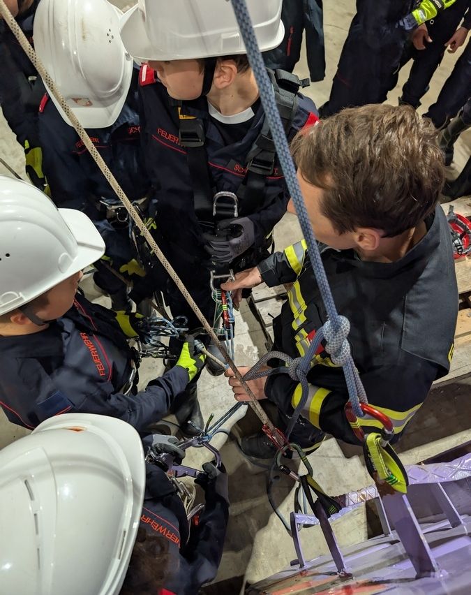 Eine Gruppe von Kindern in Feuerwehruniformen und Helmen klettert mit Hilfe eines Mannes in einem Gurtzeug eine Seilleiter hinauf.