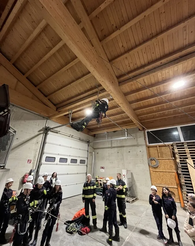 In einer Ausbildungsstätte hängt ein Feuerwehrmann an einem Seil, während Auszubildende genau zuschauen, alle tragen Helme und Schutzausrüstung, in einem Raum mit Holzbalken und einer Garagentür.