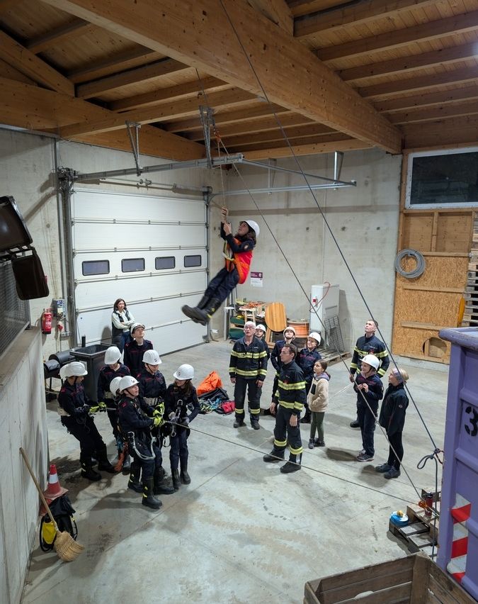 Eine Gruppe von Menschen in Feuerwehruniformen beobachtet einen Kollegen, der in einer Garage hochklettert. Der Raum hat Holzbalken, eine weiße Tür und verschiedene Ausrüstungsgegenstände.