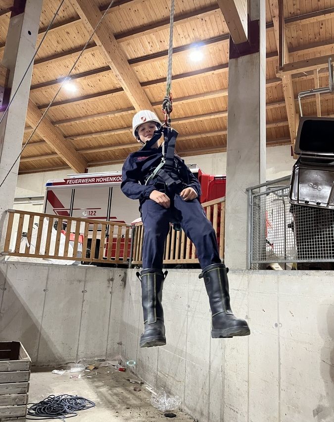 Eine Person in einer Feuerwehruniform hängt an einem Seil und trägt einen Helm. Die Szene spielt in einer Feuerwache, wobei ein Feuerwehrwagen im Hintergrund zu sehen ist.