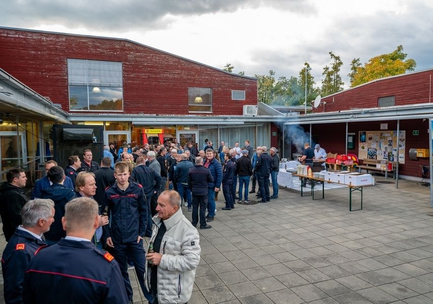 Eine Gruppe von Menschen steht vor einem roten Gebäude bei bewölktem Himmel. Einige tragen Uniformen, andere sind lässig gekleidet. Ein Tisch ist mit verschiedenen Gegenständen bestückt, und ein Mann steht in der Nähe.