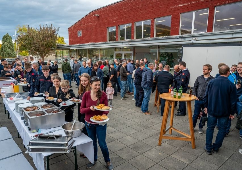 Eine Menschenmenge versammelt sich im Freien, ein Essensbuffet vor einem roten Gebäude mit Glasfenstern. Eine Frau hält einen Teller mit Essen.