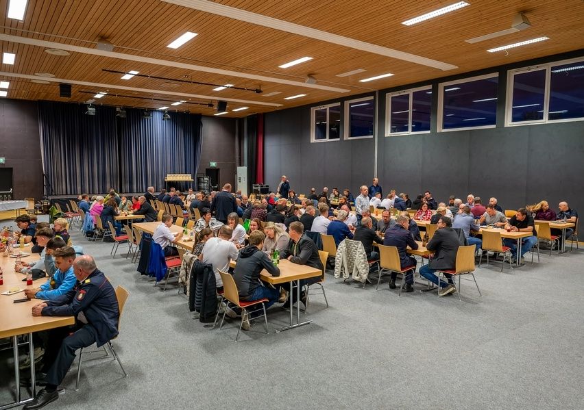 Eine große Gruppe von Menschen sitzt an Tischen in einem Konferenzraum, einige in Uniformen. Der Raum hat eine Holzdecke, große Fenster und eine Bühne mit Vorhängen. Es gibt auch Tische mit Jacken und Taschen.