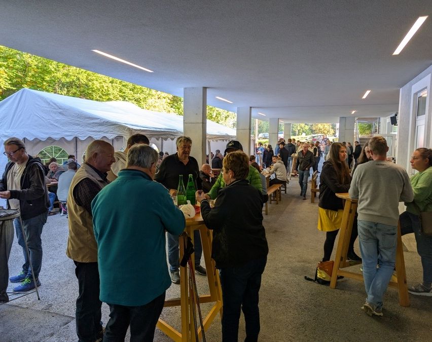 Eine Outdoor-Veranstaltung mit mehreren versammelten Personen, einige stehend und andere sitzend an Tischen mit grünen Flaschen. Im Hintergrund sind weiße Zelte zu sehen, und der Bereich befindet sich unter einem Dach.