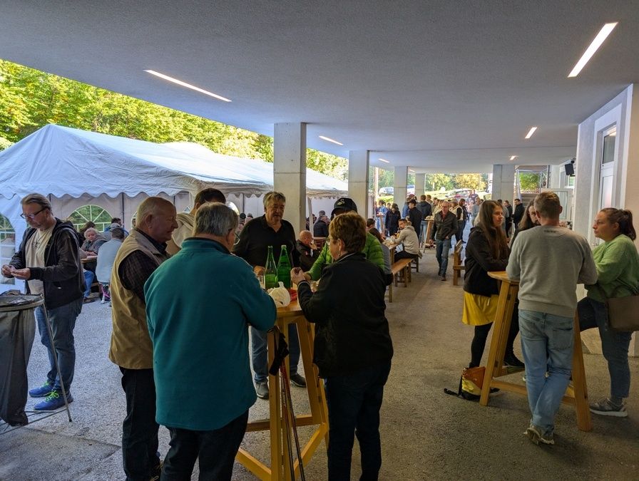 Eine Outdoor-Veranstaltung mit mehreren versammelten Personen, einige stehend und andere sitzend an Tischen mit grünen Flaschen. Im Hintergrund sind weiße Zelte zu sehen, und der Bereich befindet sich unter einem Dach.