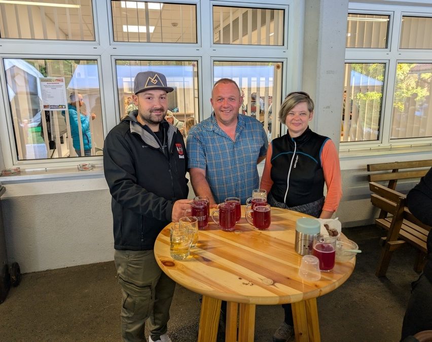 Drei Personen stehen um einen Tisch mit roten Getränken in Tassen. Der Mann links trägt einen Hut. Der Mann in der Mitte trägt ein blaues Hemd. Die Frau rechts trägt einen schwarzen Weste.
