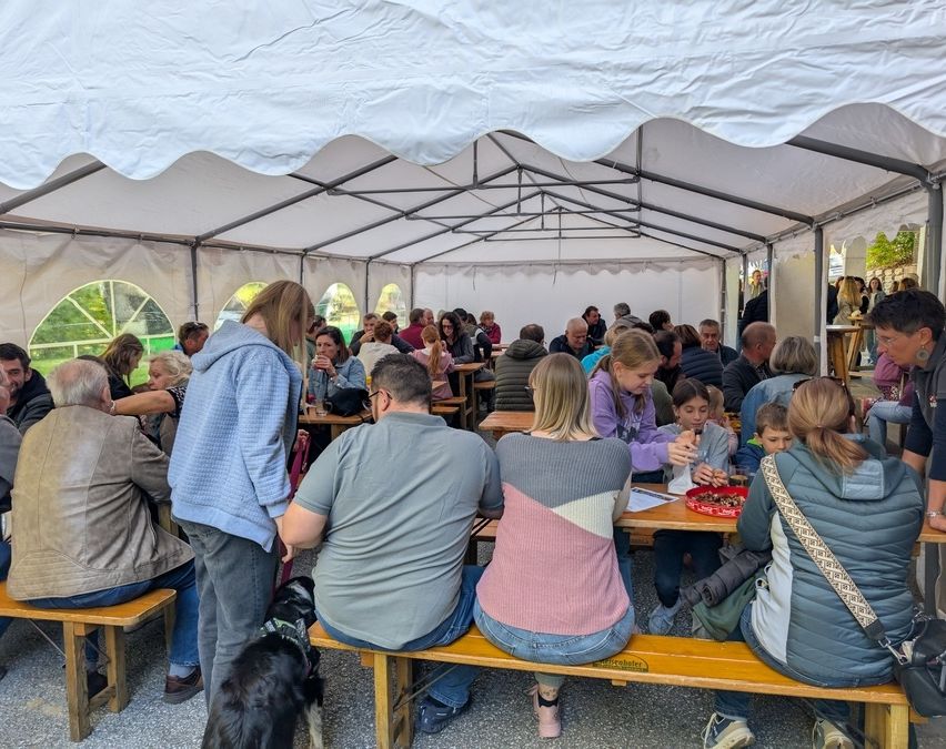 Eine Gruppe von Menschen sitzt unter einem weißen Zelt an Holztischen und genießt Essen und Getränke. Ein schwarzer Hund ist angeleint.