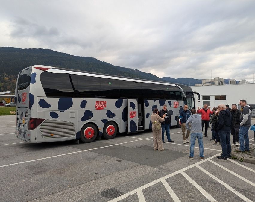 Eine Gruppe von Menschen steht in einem Parkplatz mit einem Bus mit Kuh-Muster, geöffnet, Berge im Hintergrund, bewölkter Himmel.