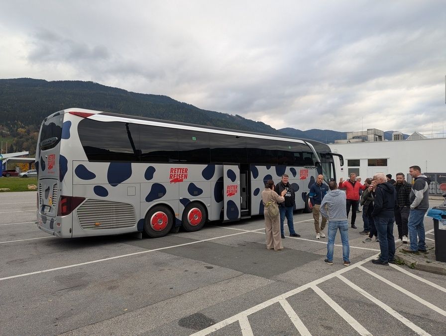 Eine Gruppe von Menschen steht in einem Parkplatz mit einem Bus mit Kuh-Muster, geöffnet, Berge im Hintergrund, bewölkter Himmel.