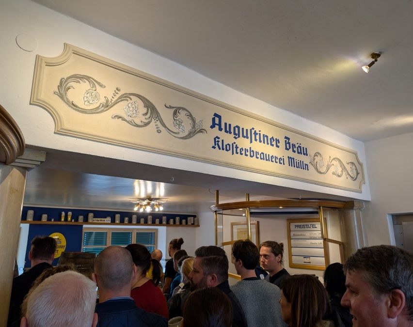 Eine Gruppe von Menschen steht in einer Bar, unter einem Schild, das 'Augustiner Beau Klosterbrauerei Mühl' steht. Die Decke hat kunstvolle Leuchten und warmes Licht.